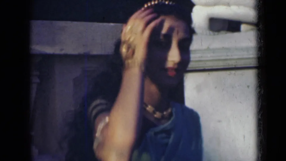 Young woman in India, 1947, lifts one hand to adjust her hair or head ornament while standing near a pale column in traditional attire.
