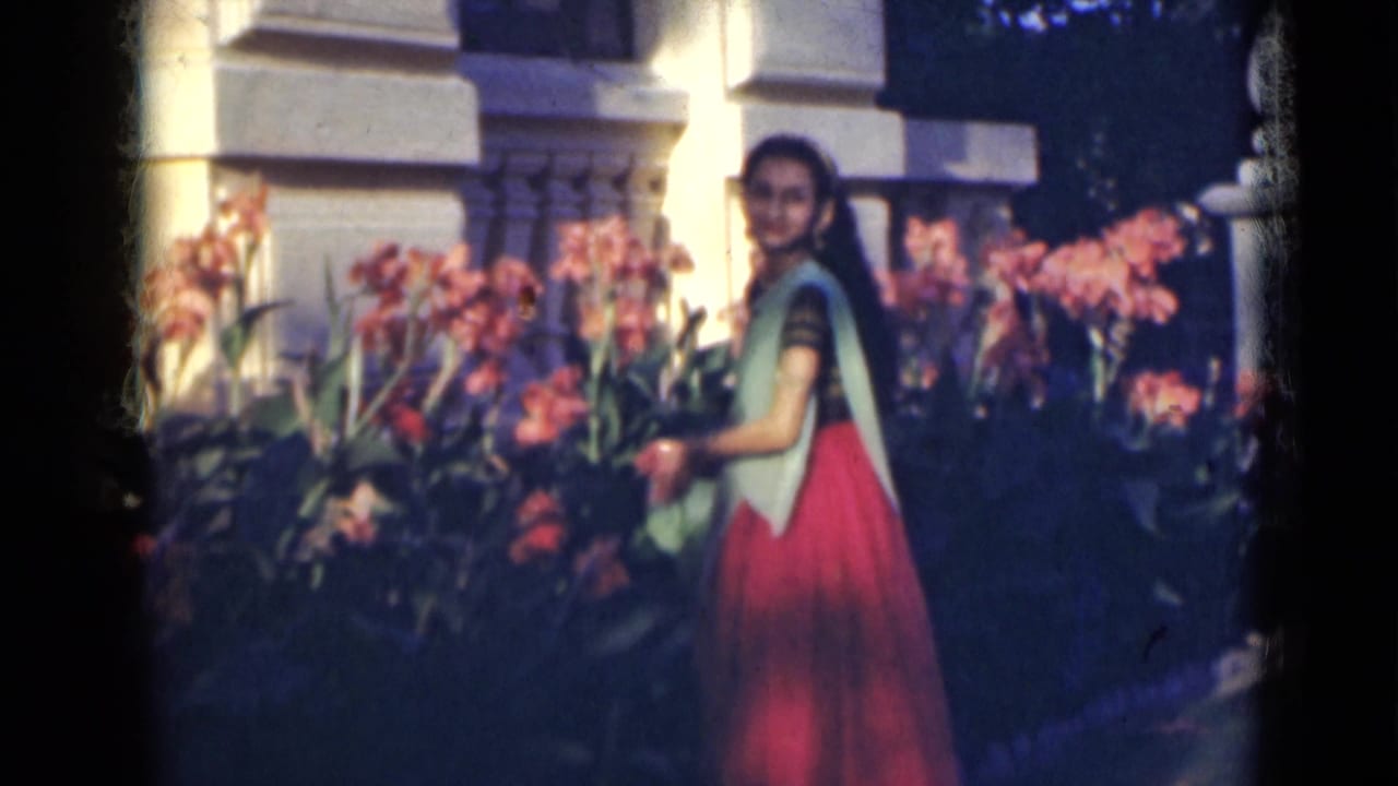 Young woman in a red and green sari stands among tall pink flowers beside white columns.