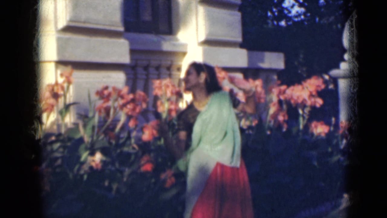 The woman in a red and green sari appears to smell or inspect the pink flowers in the 1947 India garden scene.