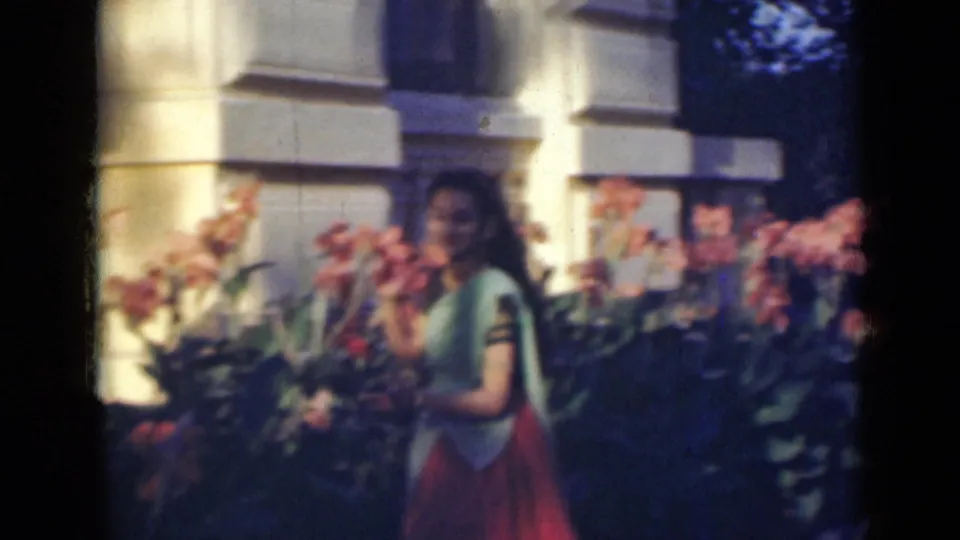 A soft 8mm frame shows the woman moving through the garden near pink flowers, with a white-columned facade behind her.