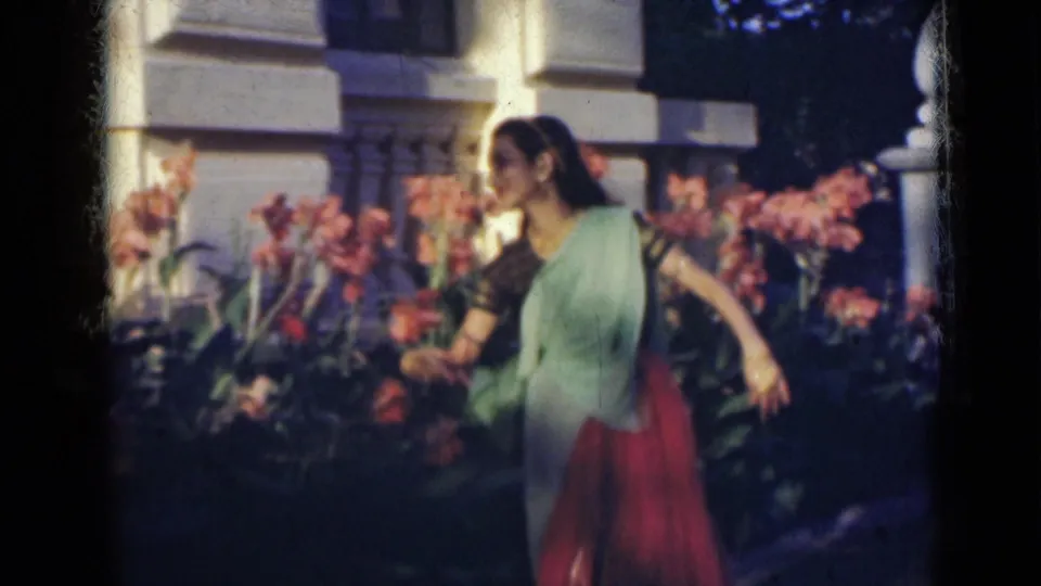 The woman extends her arms in a dance-like gesture among pink flowers in the supplied 1947 India home-movie scene.