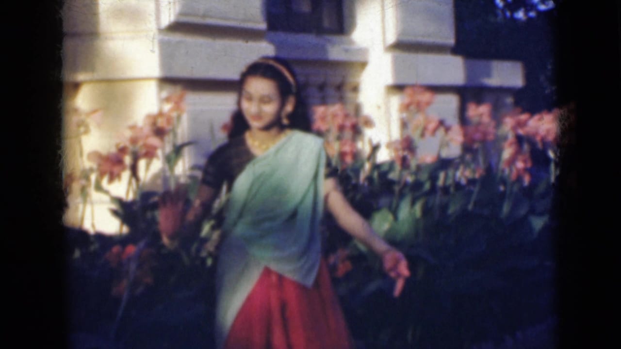 Young woman in a sari dances beside flowering plants outside a large building.