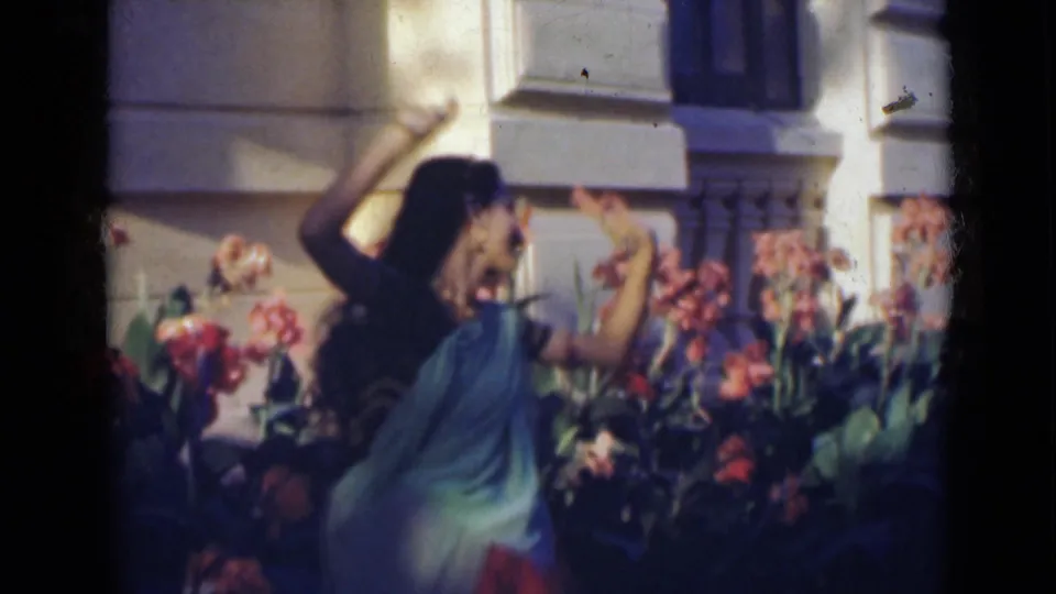 In India, 1947, the dancer turns with raised arms in front of flowering plants and the exterior wall of a large house or building.