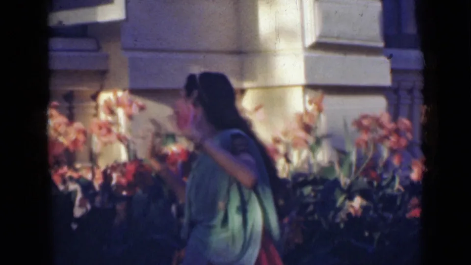 In India, 1947, the young woman is seen in profile as she dances past flowers in the garden, with a pale building facade behind her.