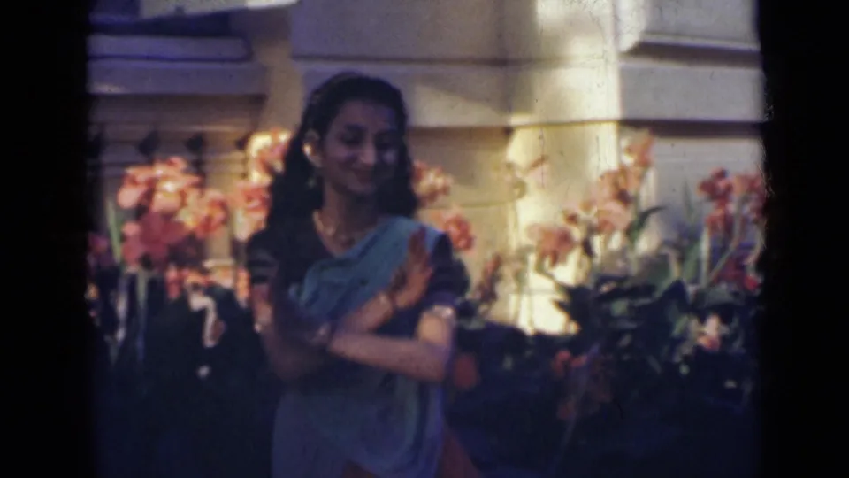 In India, 1947, the dancer faces the camera with crossed hands, framed by flowering plants and the sunlit exterior of a large building.