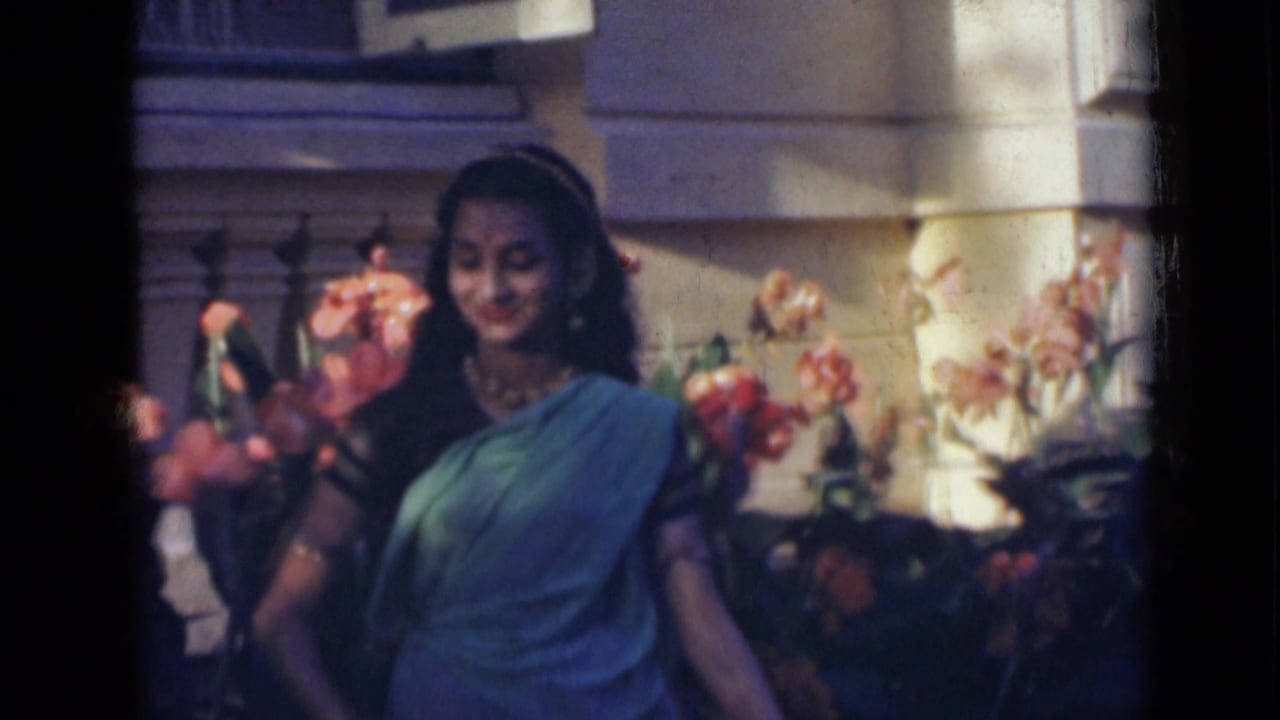 In 1947 India home-movie footage, a woman in a blue sari dances near orange flowers against a stone exterior.