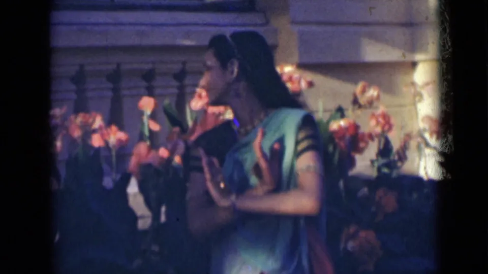 The dancer turns in profile, raising her hand in front of flowers and a stone building in metadata-supported 1947 India footage.