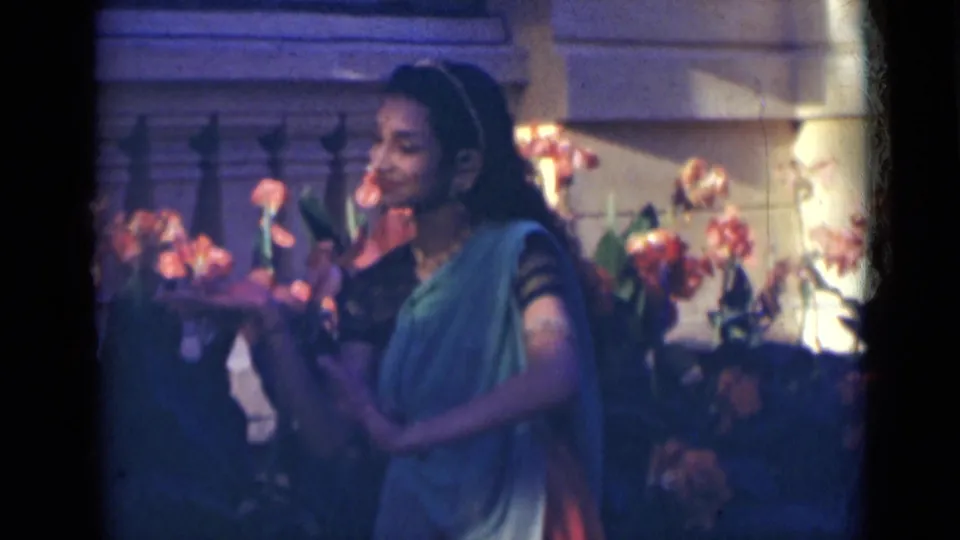 A woman in a blue sari continues a dance gesture in a garden, with flowers and exterior stonework behind her.