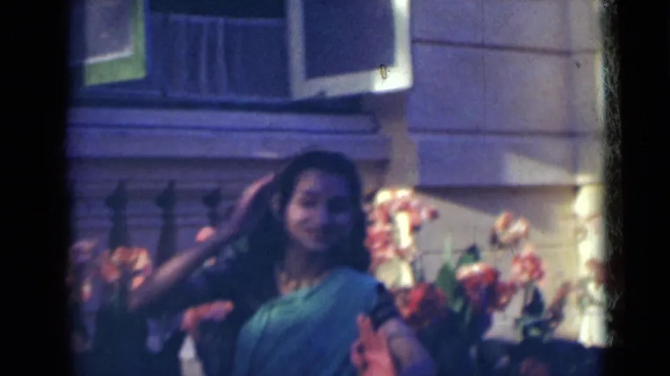 The woman raises a hand near her hair while dancing, with flowers, stone walls, and open shutters visible behind her.