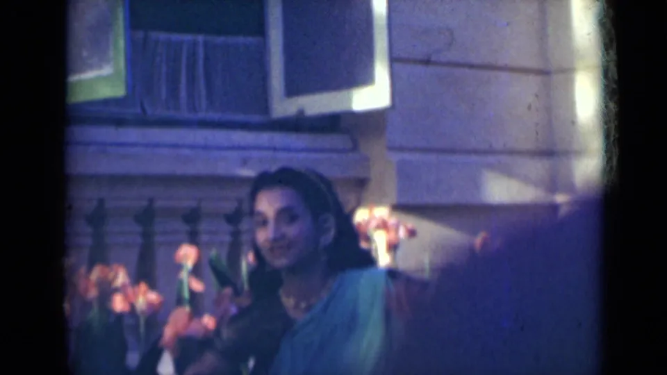 In India home-movie footage from 1947, a woman in teal clothing and jewelry dances beside orange flowers and a stone railing.