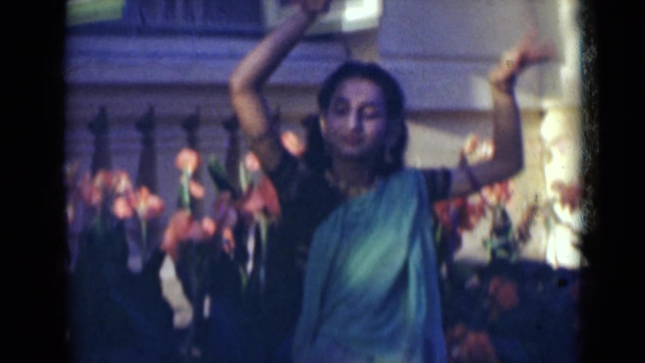 A woman in teal dress and jewelry raises one arm while dancing in 1947 India home-movie footage.