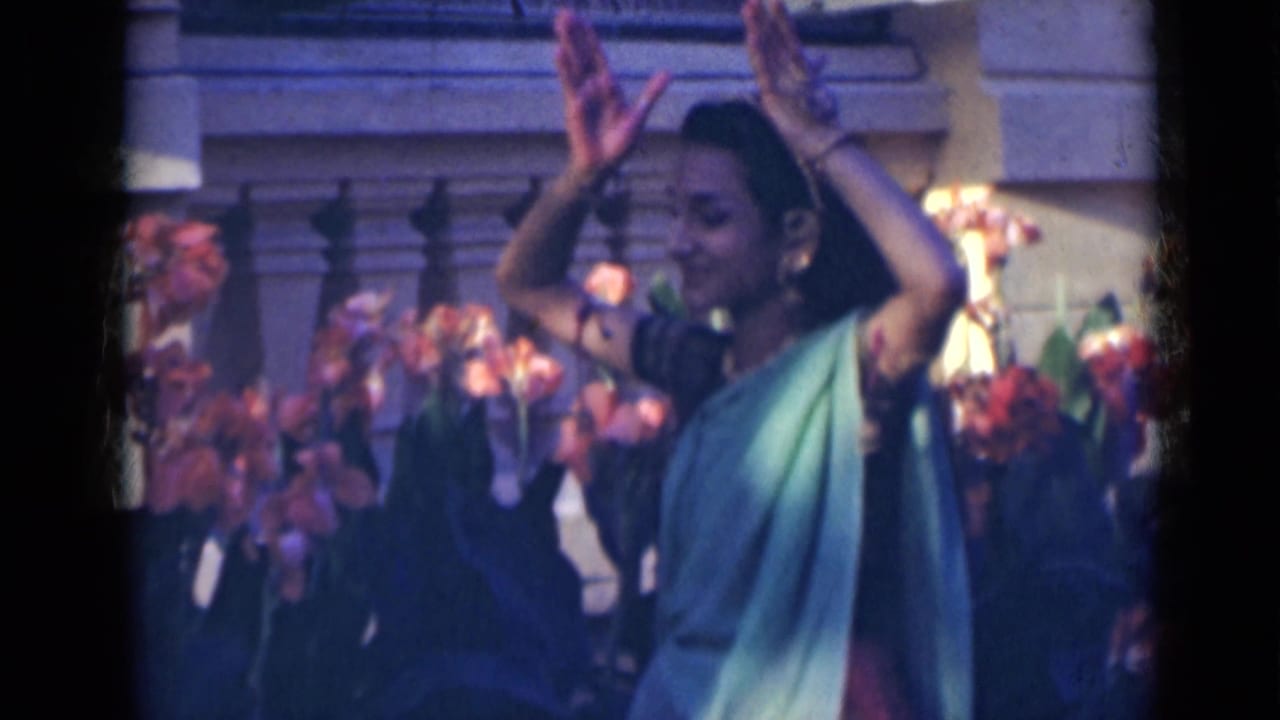 In 1947 India, a woman in a green sari performs with raised hand gestures in front of flowers and a stone balustrade.