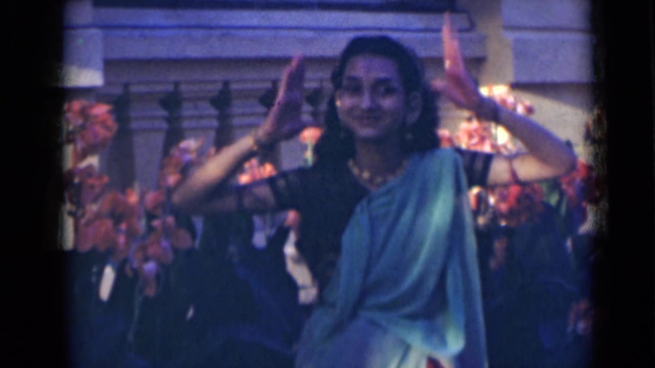 In 1947 India, the dancer faces the camera with lifted hands, jewelry, and a green sari against a decorated stone setting.