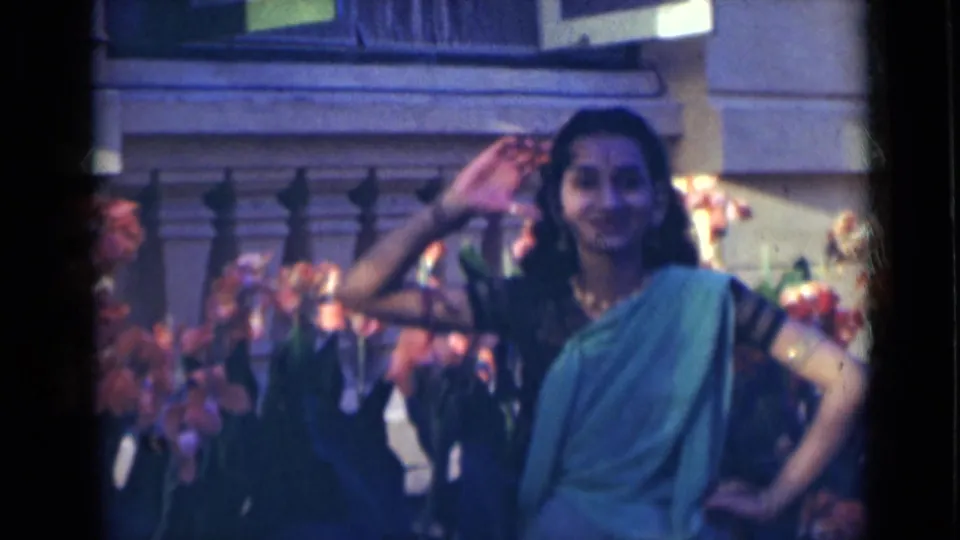 In 1947 India, the performer holds a poised dance stance with one hand raised and the other at her waist before flowers.