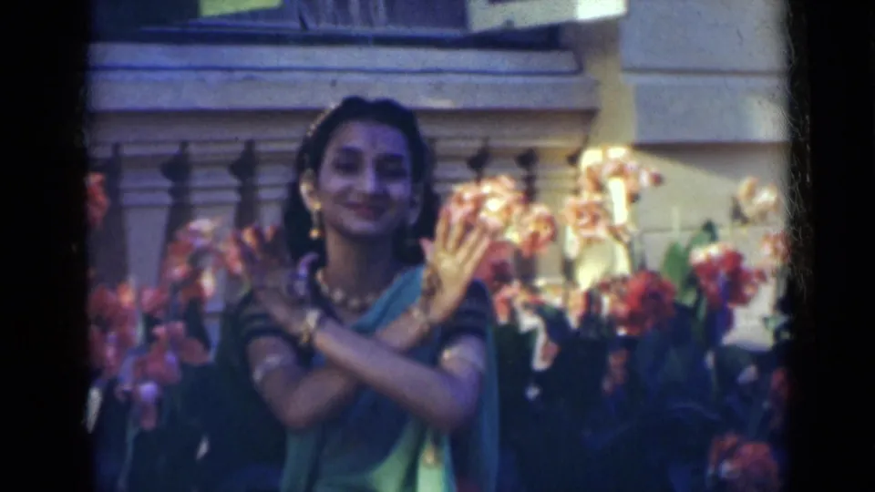 In 1947 India, the dancer smiles toward the camera with crossed hands, jewelry, flowers, and a stone balustrade behind her.