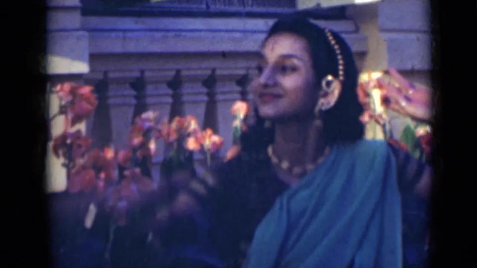 In a soft 8mm frame, the young woman smiles slightly while dancing in a green sari near blooming plants.