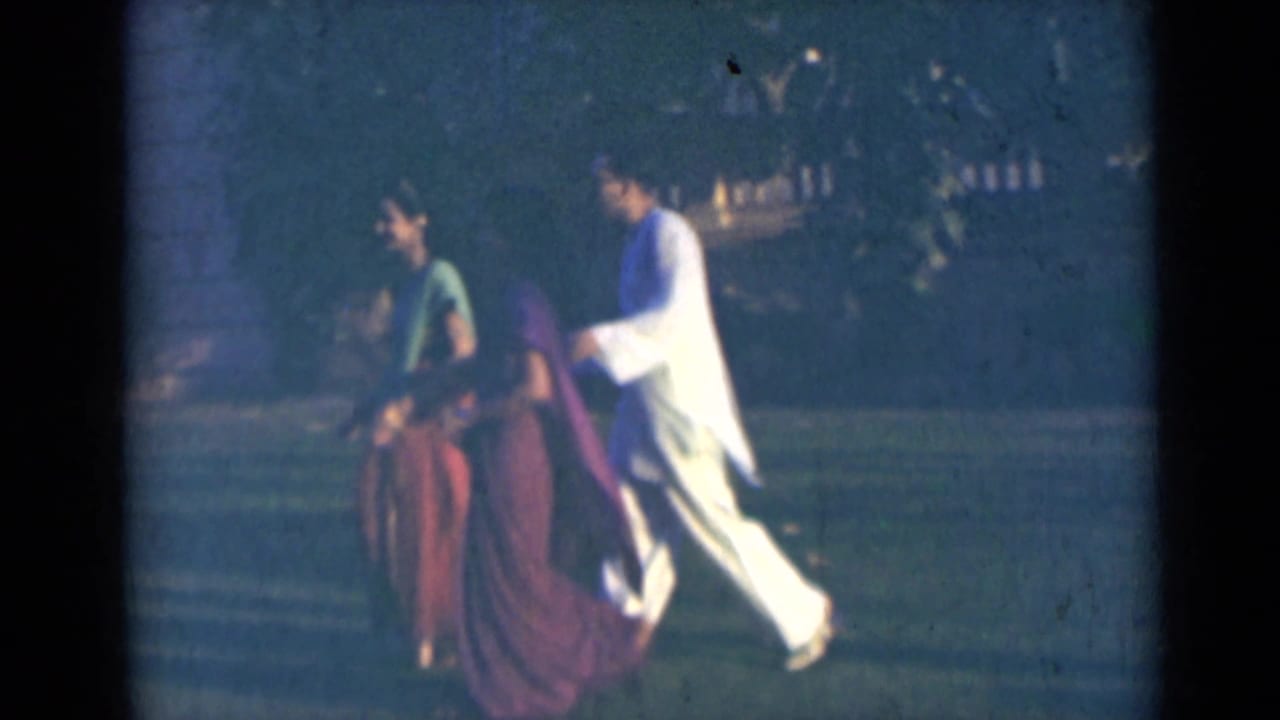 In India 1947 home-movie footage, adults in sarees and light kurtas walk left to right outdoors near trees and a building.