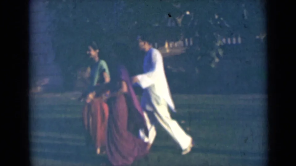 In India 1947 home-movie footage, adults in sarees and light kurtas walk left to right outdoors near trees and a building.
