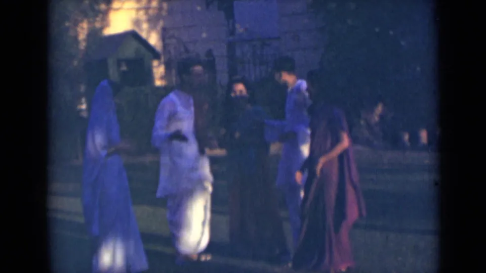 Five adults in India 1947 home-movie footage stand and meet outdoors near trees and a building, with visible film grain and low light.