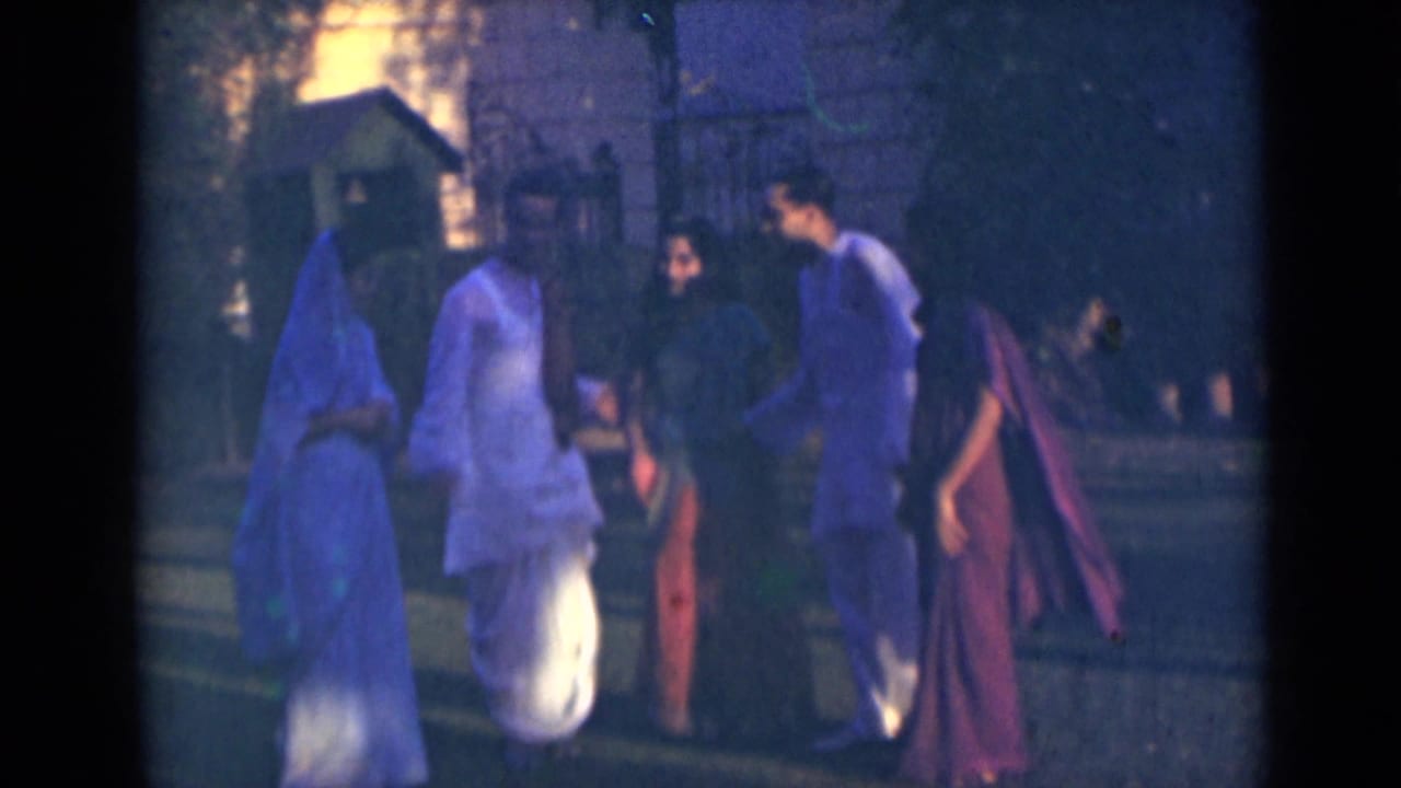 Five adults walk together outdoors, wearing light and red draped clothing near steps and foliage.