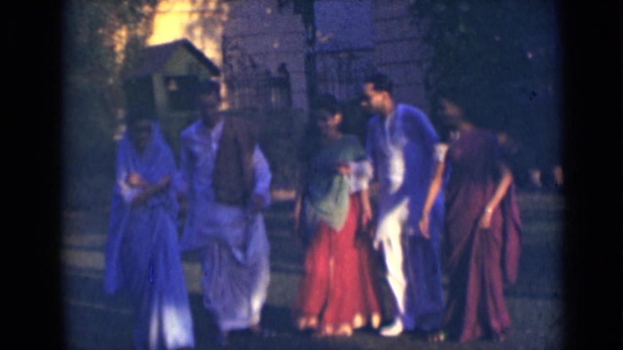 Five adults in India walk together across a garden-like area, with saree-like garments and light kurtas visible.