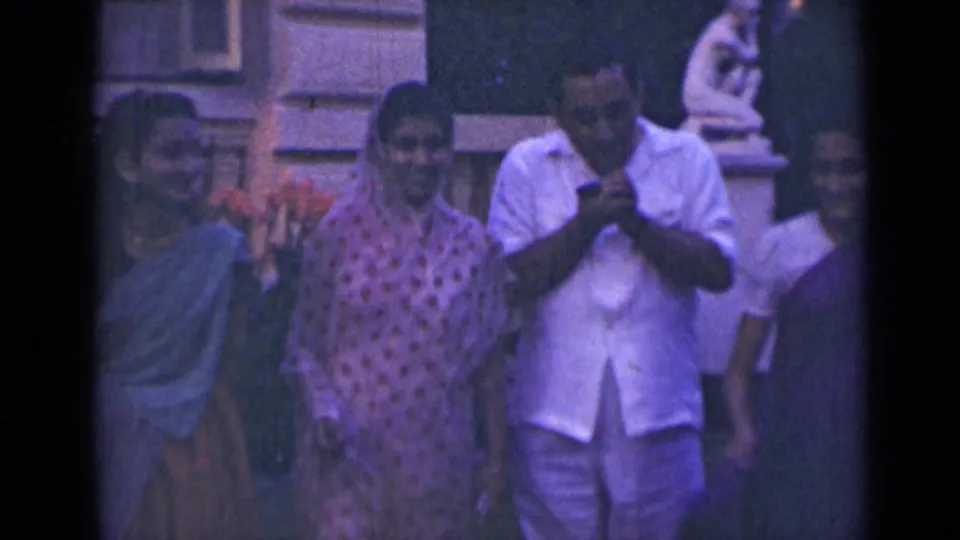 The group remains outside in 1947 India, with women in traditional attire watching as the man handles a cigar-like object.