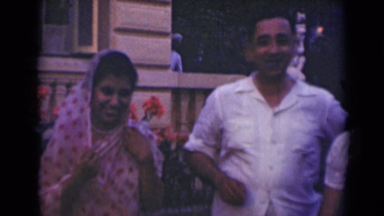 1947 India home-movie frame with two adults smiling outdoors beside red flowers and a formal stone building facade.