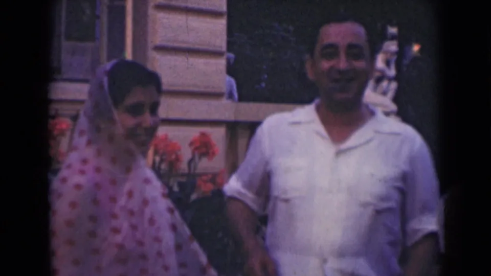 1947 India 8mm frame of a woman in a pink patterned covering turning near a smiling man outside a stone building.