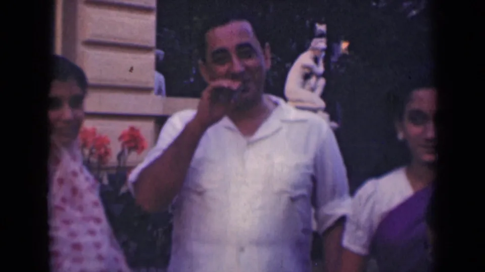 1947 India home-movie frame showing a smiling man gesturing near his mouth with two women partly visible beside him.