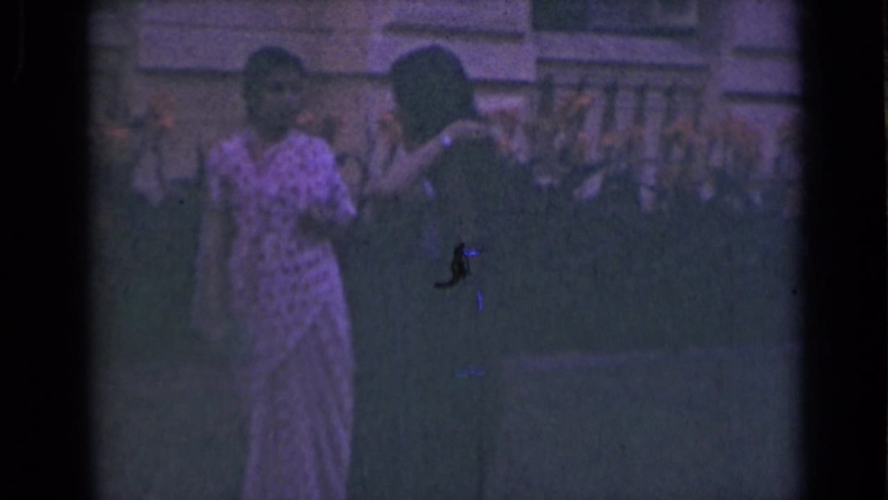 Two women in saris appear outdoors in 1947 India, standing near a building facade and balustrade in dim 8mm color.