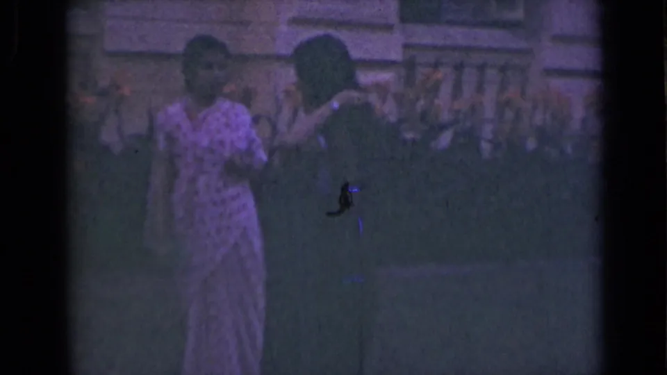 Two women in saris appear outdoors in 1947 India, standing near a building facade and balustrade in dim 8mm color.