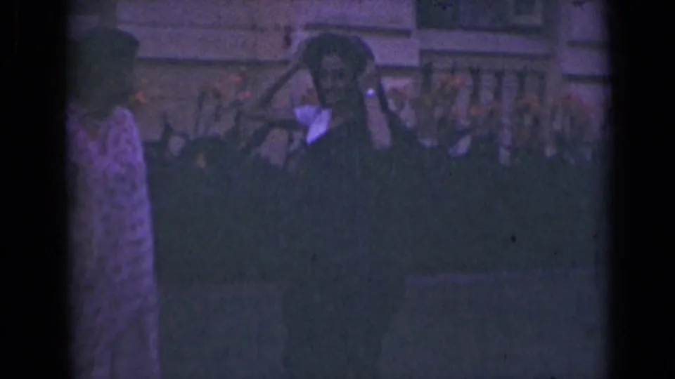 One woman appears to raise her hands toward another woman's head covering, with a balustrade and building behind them.