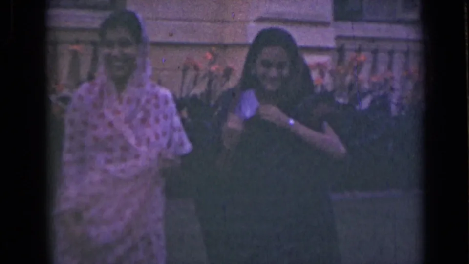 Two women in saris stand together and smile toward the camera in an outdoor 1947 India home-movie frame.