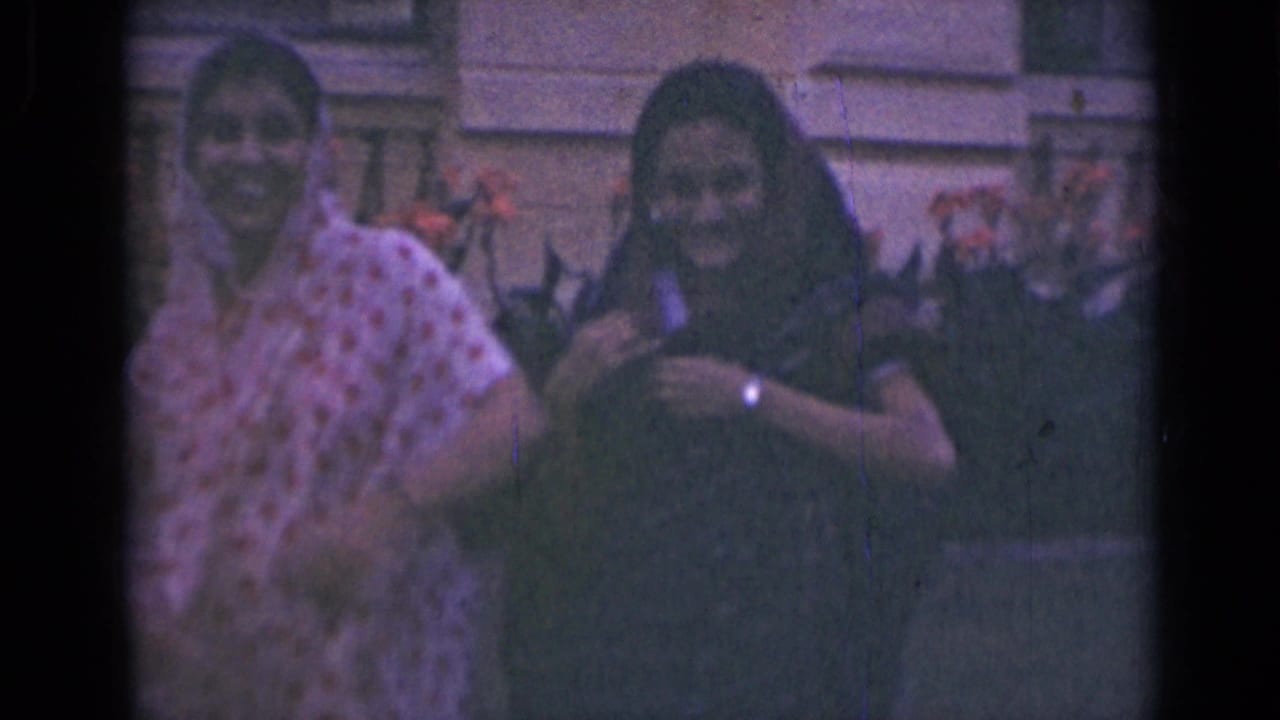 Two women in sarees smile toward the camera in archival home-movie footage from India, with soft focus and faded color.