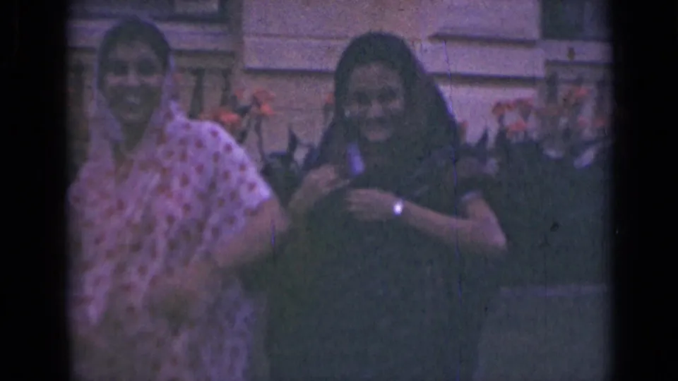Two women in sarees smile toward the camera in archival home-movie footage from India, with soft focus and faded color.