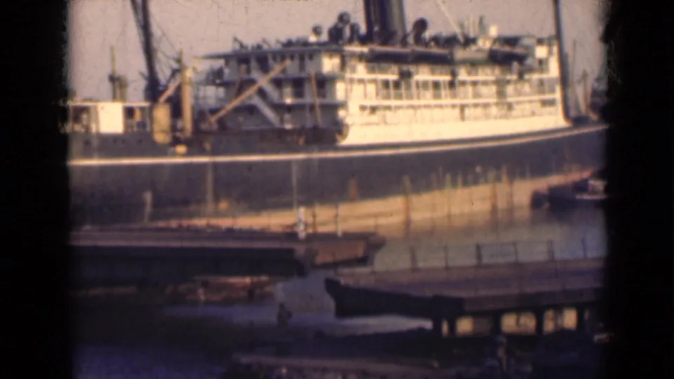 The moored steamship is filmed from shore, with water, dock structures, and a small boat visible in the foreground.