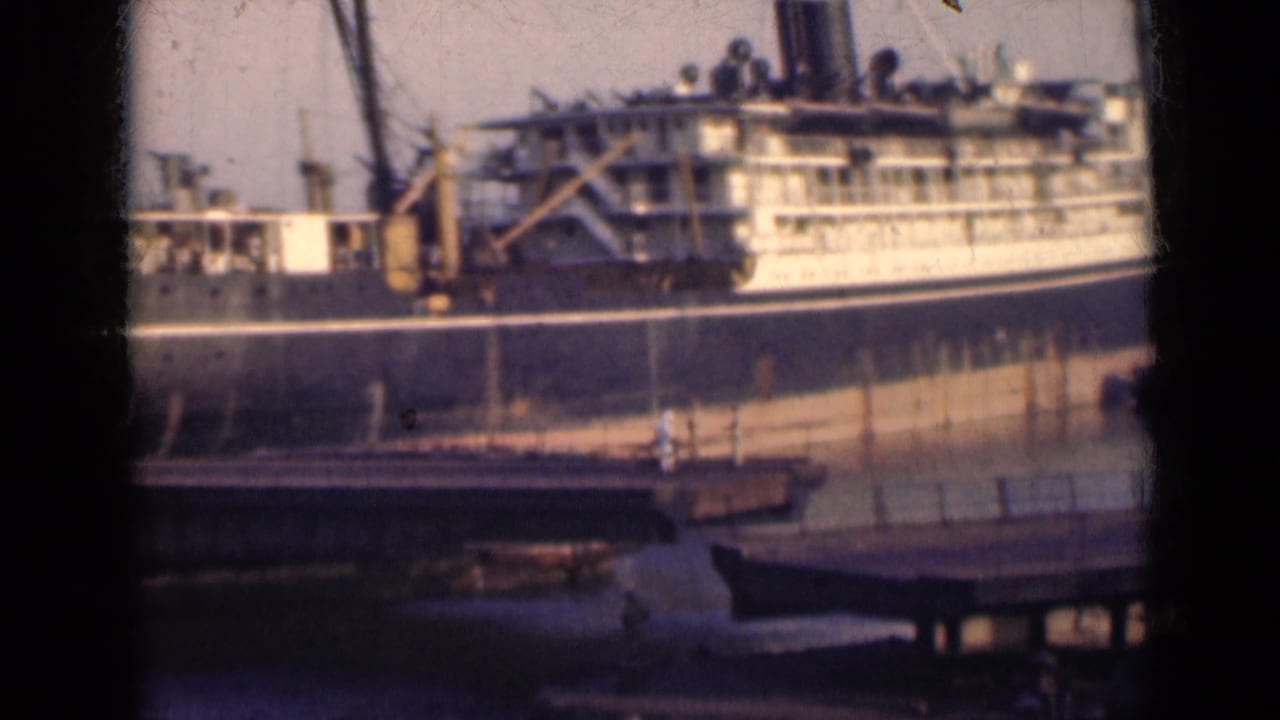 A dark-hulled passenger ship with white upper decks sits at an Indian port in 1947, seen beyond a bridge in soft 8mm color.