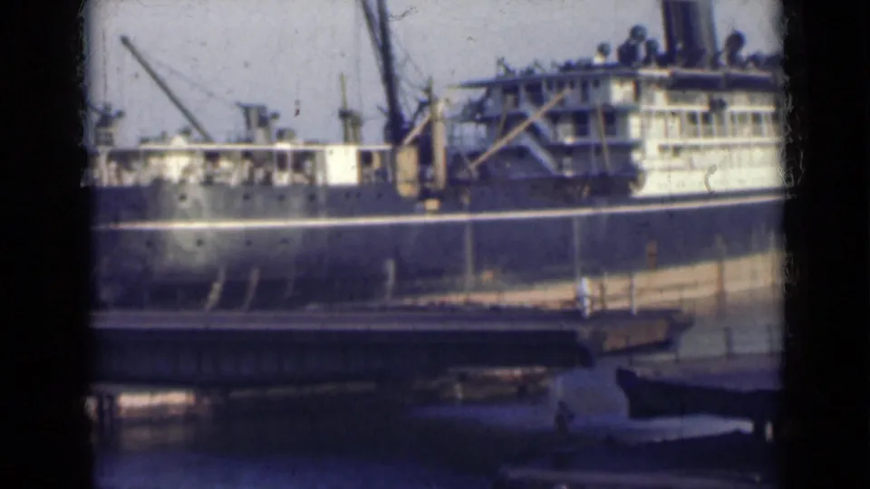 The docked ship remains framed across the water, with a bridge in the foreground and port structures behind it in 1947 India footage.