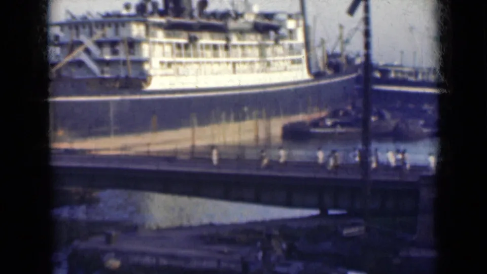 1940s India home-movie frame showing a docked passenger ship behind a bridge with people moving across the foreground walkway.