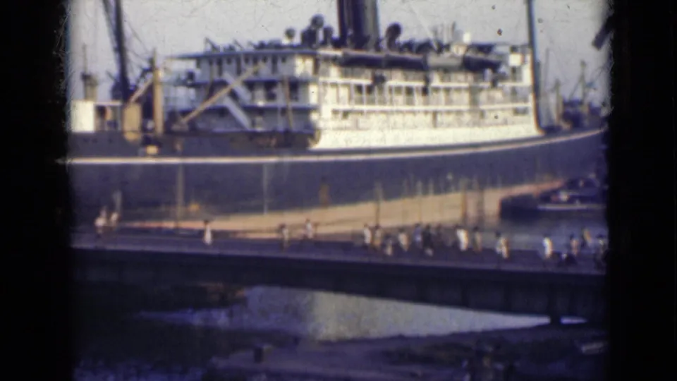 Distant 8mm view of a docked passenger ship in India with people crossing a bridge in front of the hull.