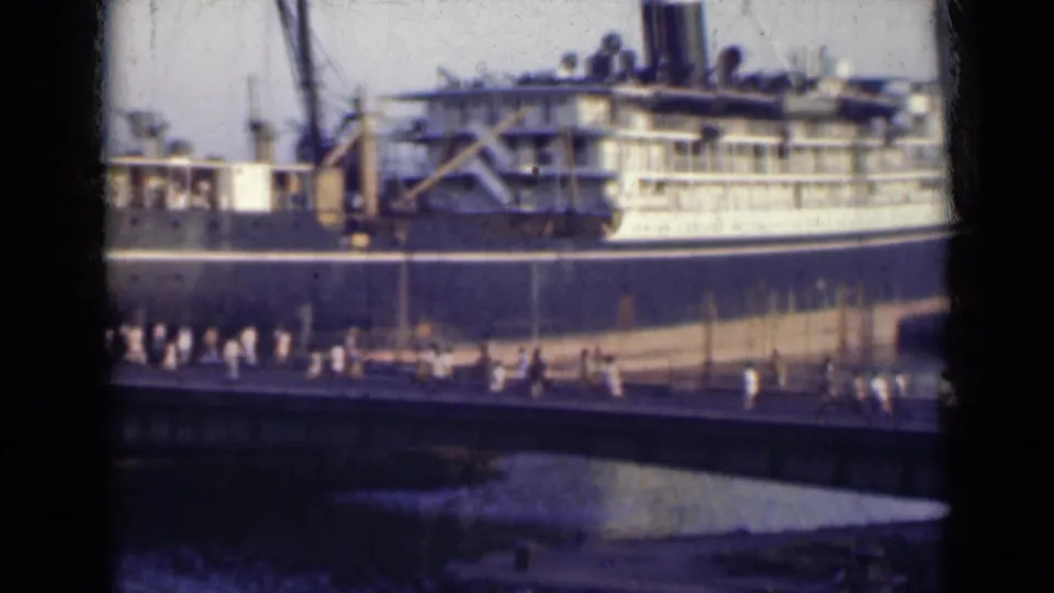 1940s India 8mm frame with a docked passenger ship, visible upper decks, and a clustered line of people on the bridge below.
