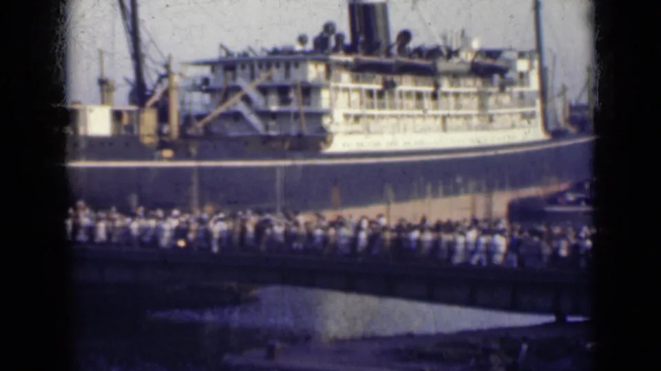 1947 India home-movie frame showing a dense crowd crossing a dockside bridge toward a large ocean liner.