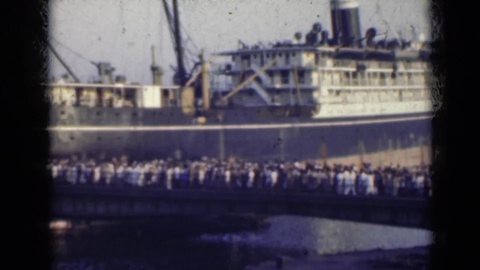 1947 India home-movie frame showing a distant crowd crossing toward or beside a large passenger ship at port.