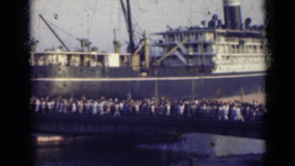 1947 India home-movie frame showing people moving across a bridge in front of a dark-hulled passenger ship.