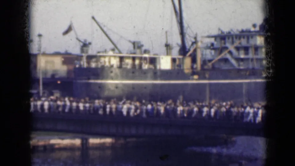 Home-movie view of people on a waterfront bridge with a docked ship, cranes, and a small flag in the background.