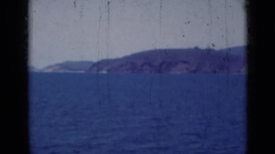 A 1947 India coastal view shows a distant hilly shoreline across blue water, with dust, scratches, and soft 8mm color visible.