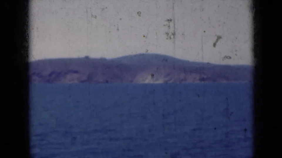 1947 India 8mm frame of blue ocean and a distant mountainous coastline, with faded color, dust marks, and soft focus.