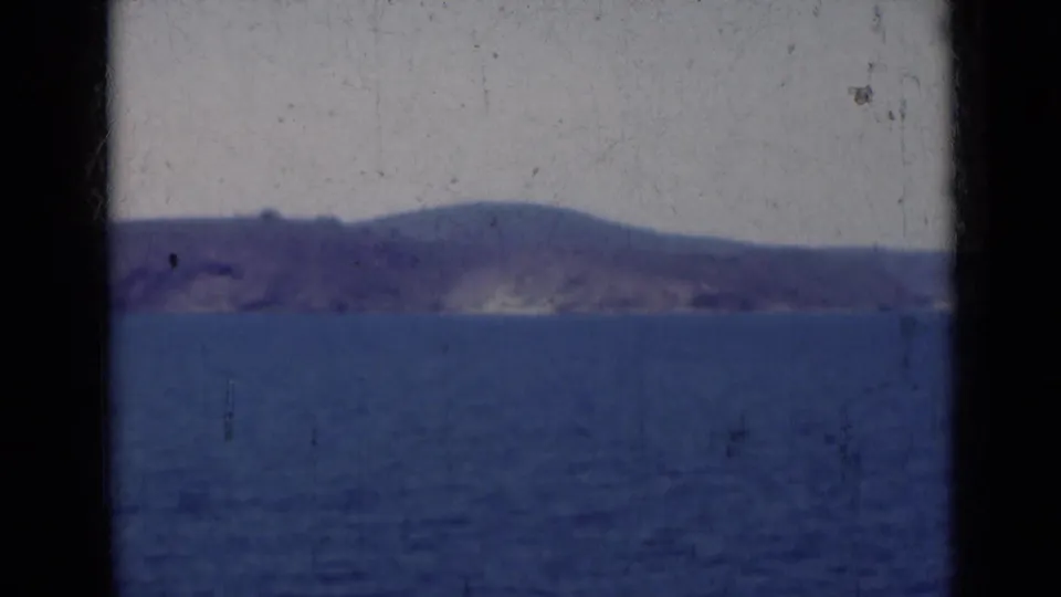 1947 India 8mm frame showing calm blue water and a hazy shoreline or island in the distance beneath a pale sky.