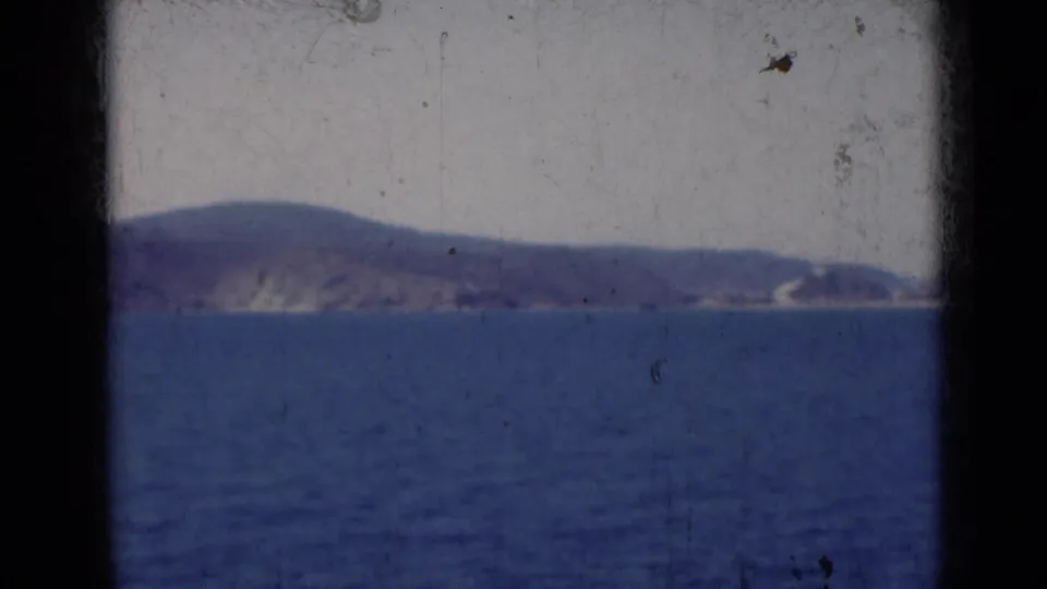 1947 India home-movie frame showing a soft, distant shoreline beyond blue water, with aged 8mm surface marks visible.