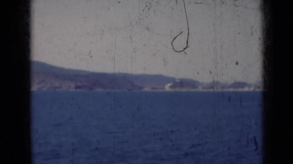 1947 India 8mm frame showing open blue water, a distant hazy shoreline, and low hills beneath a pale sky.
